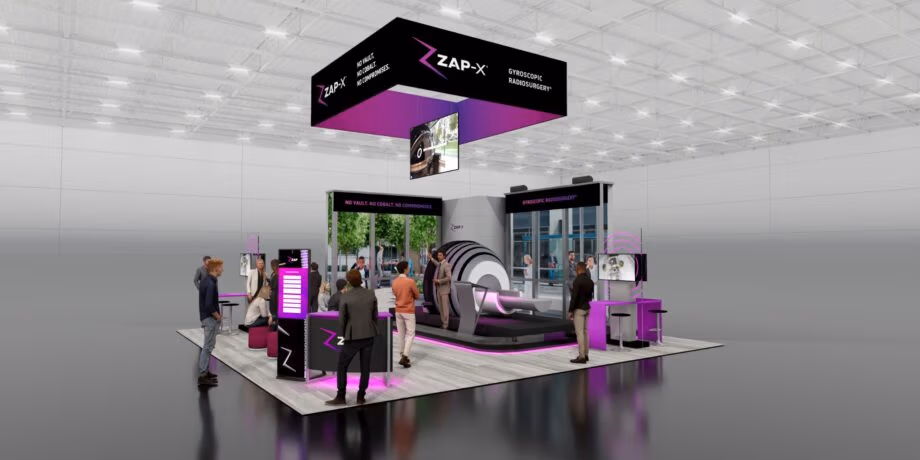 Exhibit booth for ZAP Surgical at ASTRO 2025 showcasing the debut of the ZAP-X® Gyroscopic Radiosurgery® system. The booth features bold black and purple branding with illuminated signage reading “No Vault. No Cobalt. No Compromises.” A suspended overhead cube displays the ZAP-X logo, while large digital screens present video demonstrations and clinical images. At the center, a display of the ZAP-X treatment system is highlighted with purple lighting accents. The booth includes a reception counter with monitors, seating for attendees, and modern architectural panels featuring the tagline “Brain Radiosurgery deserves better than ‘good enough.’ DEMAND EXCELLENCE.”