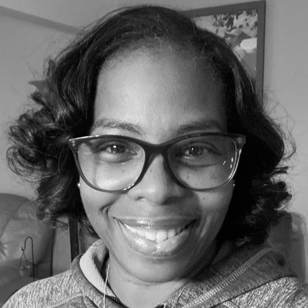 A smiling ZAP-X patient named Antoinette is shown in a close-up, black-and-white photograph. She is wearing glasses and a hoodie, with neatly curled hair. Antoinette is seated comfortably indoors, with a soft couch and framed artwork visible in the softly lit background. Her expression is warm and cheerful.