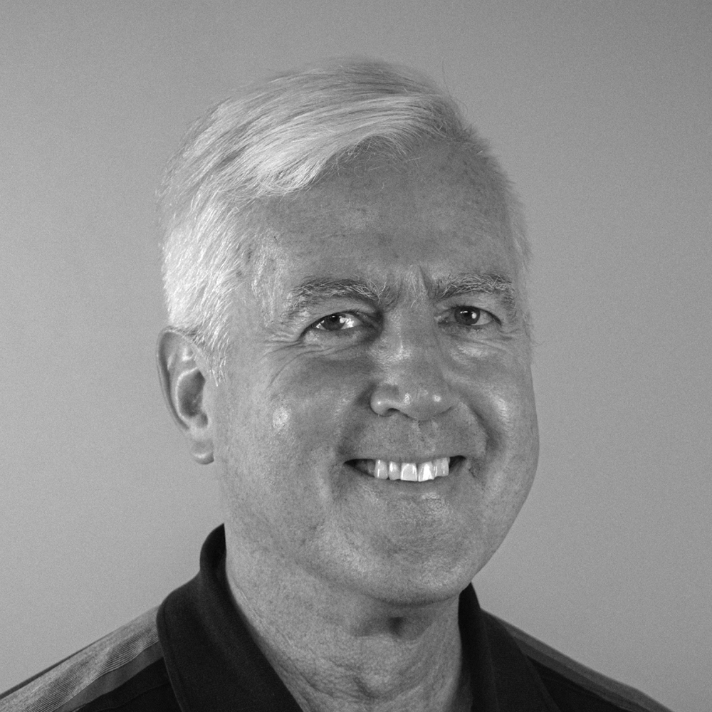 A smiling ZAP-X patient named Claus is shown in a close-up, black-and-white photograph. He has short white hair and is wearing a collared shirt. Claus appears in a well-lit, neutral background setting, and his expression is warm and friendly.