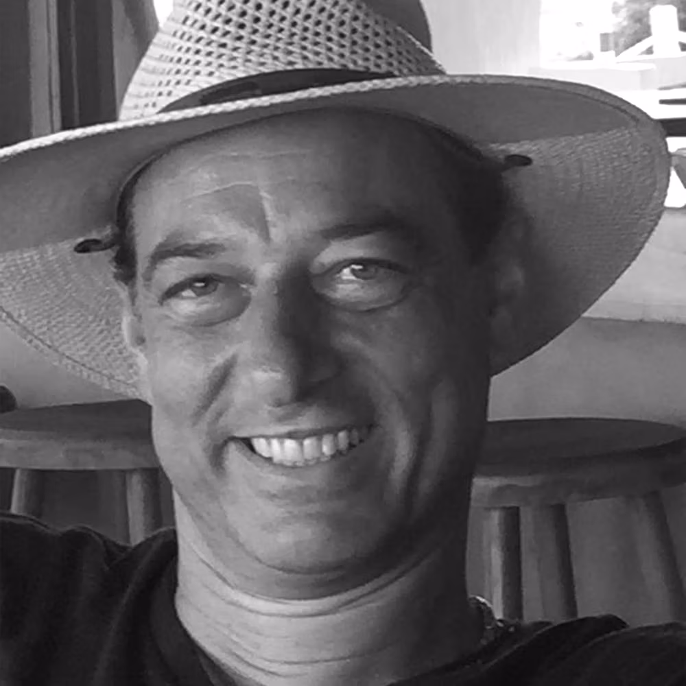 A smiling ZAP-X patient named Hagen is shown in a close-up, black-and-white photograph. He is wearing a wide-brimmed sun hat and a relaxed shirt, seated comfortably in an indoor setting with wooden stools visible in the background. Hagen’s expression is cheerful and relaxed.