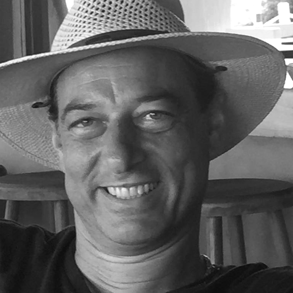 A smiling ZAP-X patient named Hagen is shown in a close-up, black-and-white photograph. He is wearing a wide-brimmed sun hat and a relaxed shirt, seated comfortably in an indoor setting with wooden stools visible in the background. Hagen’s expression is cheerful and relaxed.