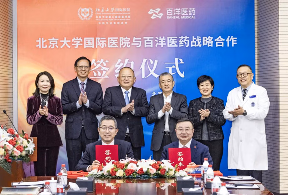 The photo shows a formal signing ceremony between Peking University International Hospital and Baheal Medical. Two representatives sit at the table in front, each holding a red folder marked “签约本” (signing document), while several other executives and medical professionals stand behind them, smiling and applauding. The background banner features both institutions’ logos and reads, “Strategic Cooperation Signing Ceremony between Peking University International Hospital and Baheal Medical.” The setting appears professional and celebratory, with flower arrangements and documents placed neatly on the conference table.